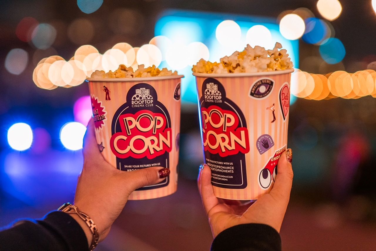 An image of people holding two containers of popcorn with Rooftop Cinema Club as the logo. 