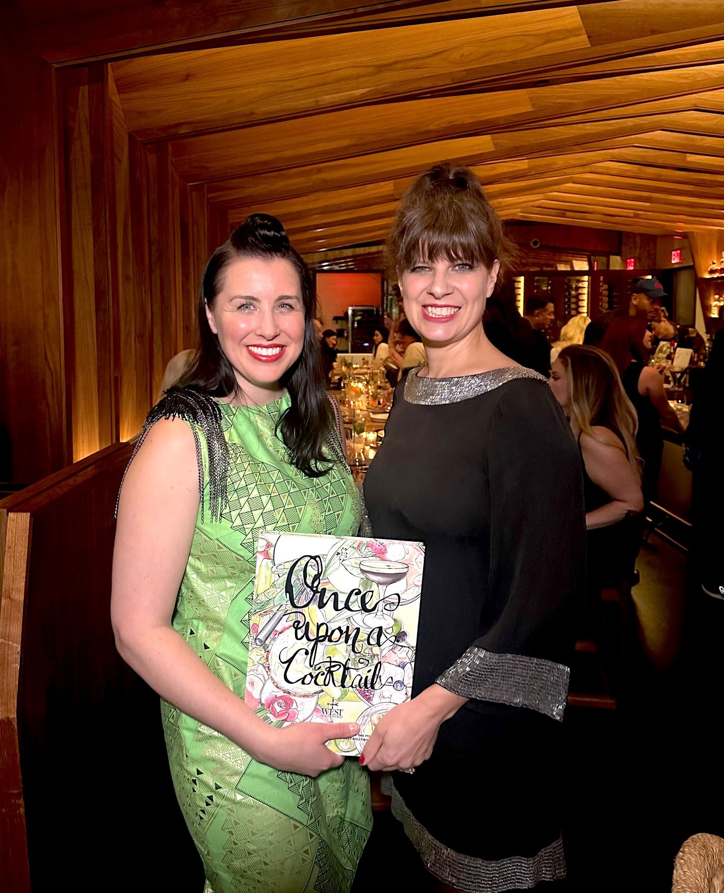 An image of Once Upon a Cocktail's Imagineer & illustrator Kate Brightside (l) and cocktail curator Sarah L.M. Mengoni (r) posing with their cocktail recipe book at their International Women's Day celebration at Soulmate.