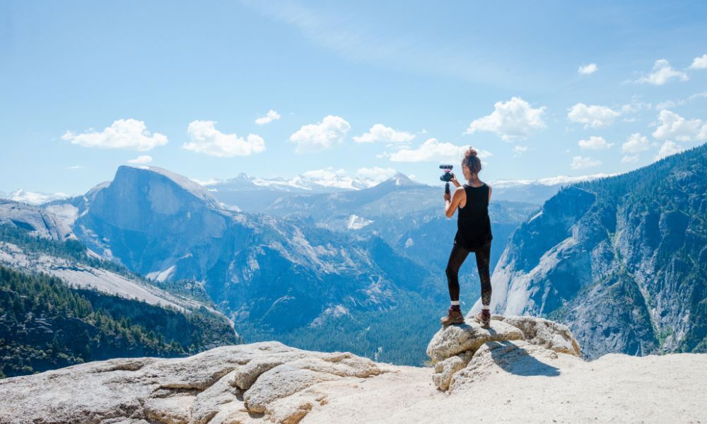 An image of a woman filming mountains from the edge of a cliff.
