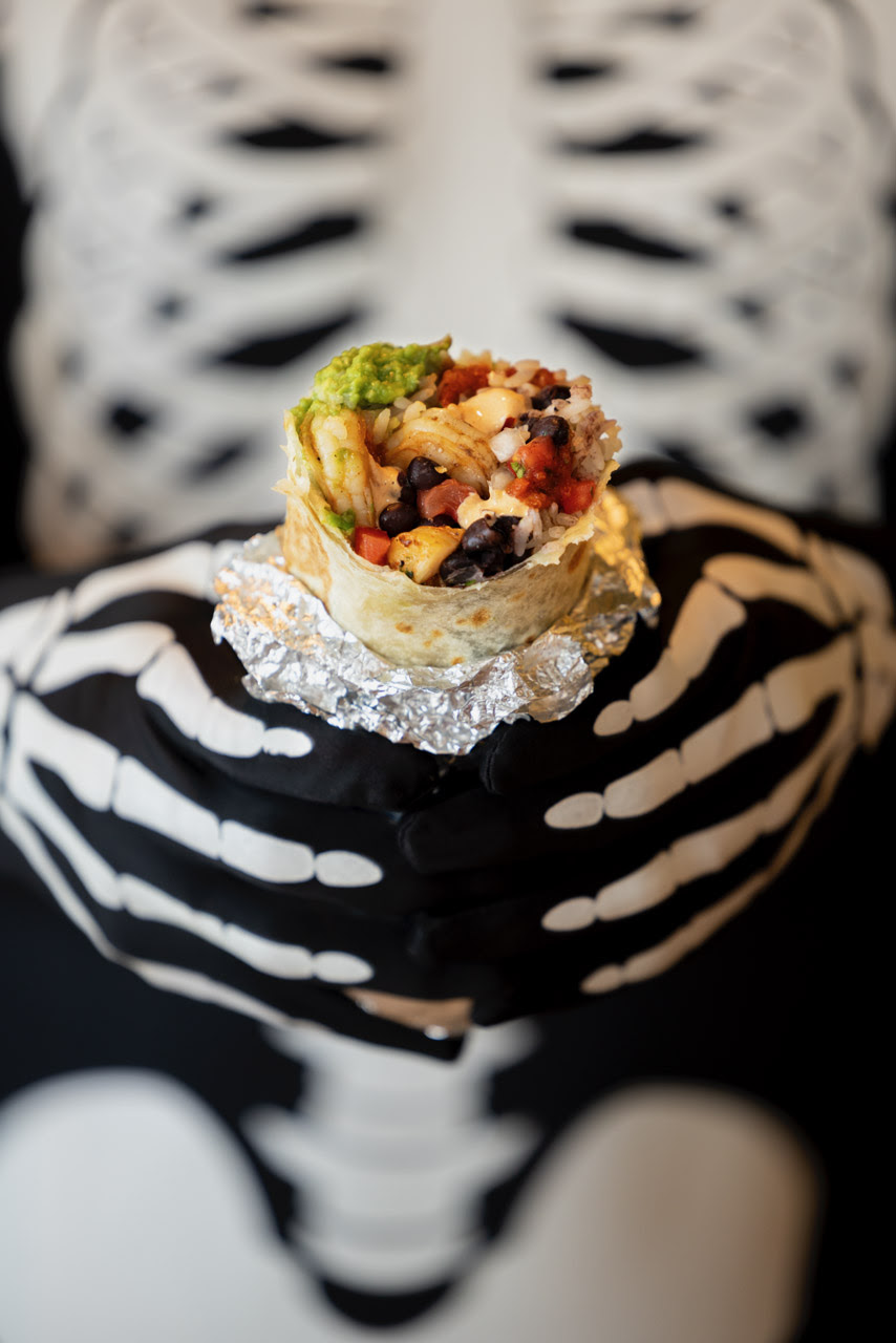 An image of someone dressed as a skeleton holding a Rubio's burrito for a Happy Halloween.