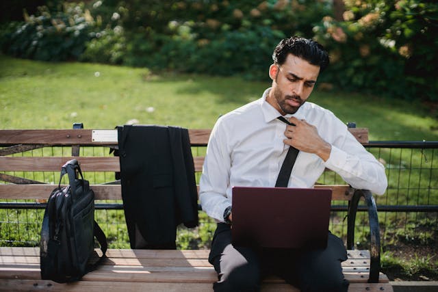 An image of a man sitting on an outdoor bench with his laptop.