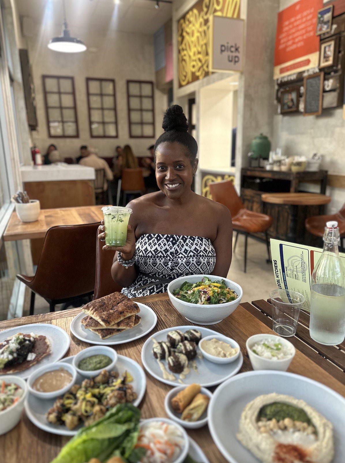 An image of lifestyle blogger Ariel holding a Persian Cucumber Mint Lemonade in front of a spread at Local Kitchens.