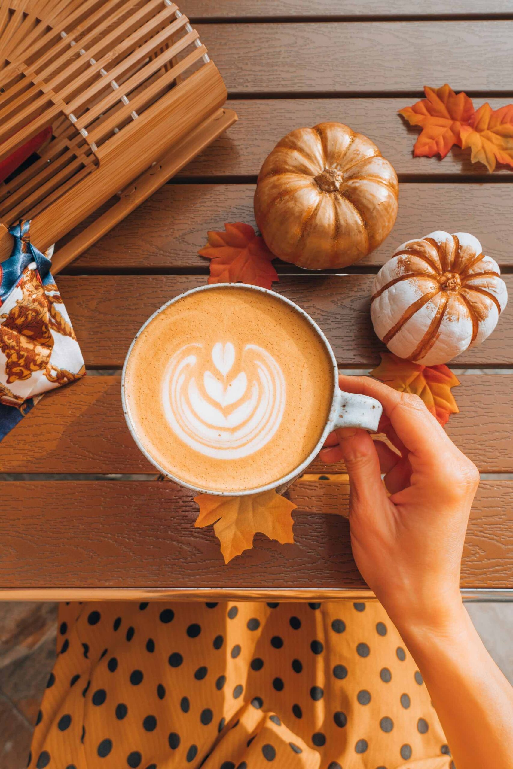 An image of someone holding a pumpkin spice latter on a table with mini pumpkins surrounding it.