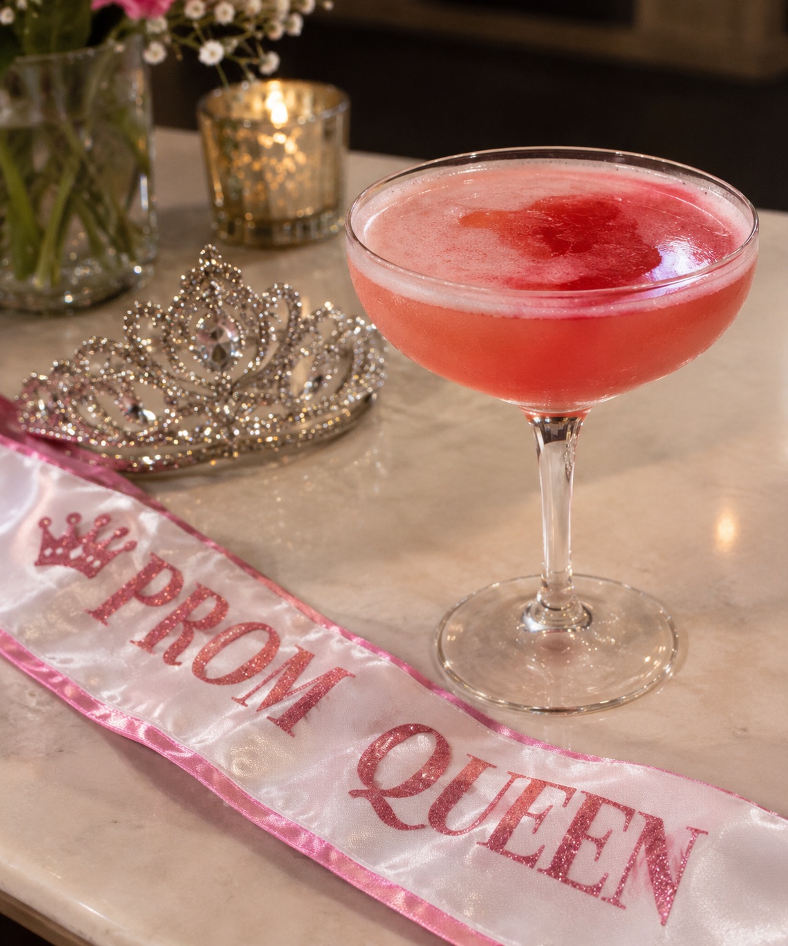 An image of a cocktail surrounded by a prom queen tiara and a prom queen sash.